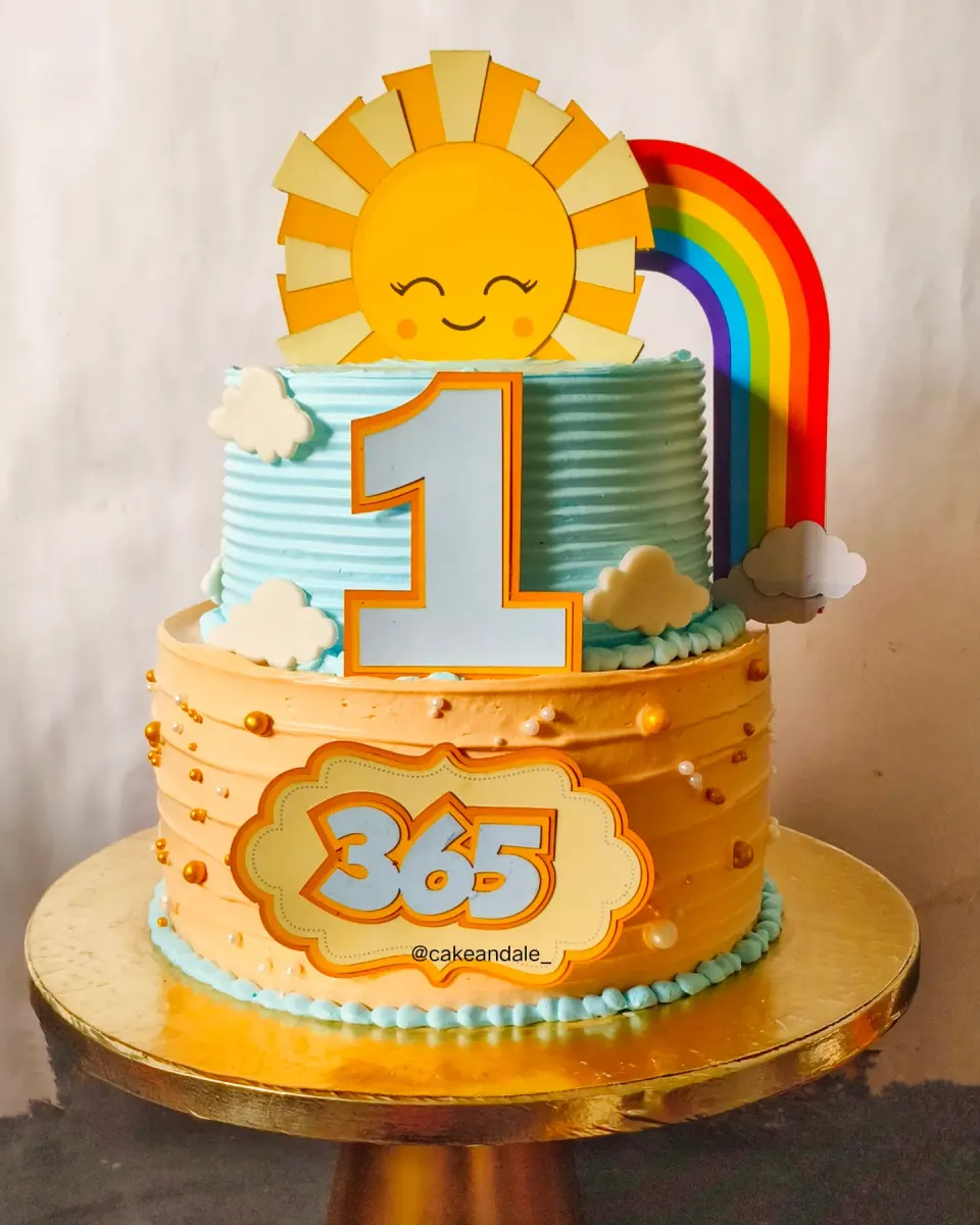 Two tier first birthday cake with a smiling sun, rainbow and clouds, celebrating 365 days