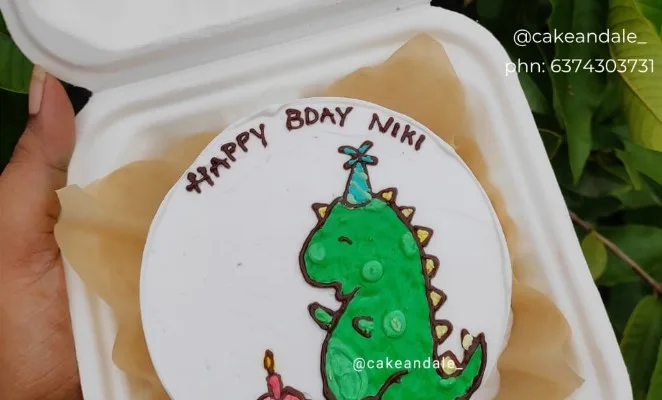 Hand-piped dinosaur bento cake in a lunch box reading Happy Bday Niki